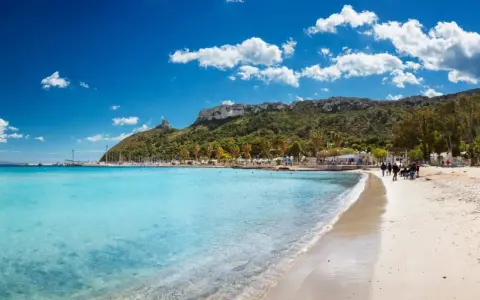 Spiaggia con acque cristalline e la sagoma della Sella del Diavolo all'orizzonte, sotto un cielo azzurro con nuvole bianche.