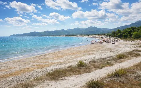 Spiaggia affollata con ombrelloni e gente che si gode il sole. Il nuoro mare azzurro lambisce la costa, con montagne verdi sullo sfondo.