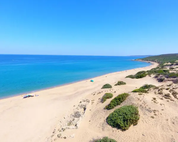 Vasta spiaggia di sabbia dorata, mare azzurro e vegetazione mediterranea sulla spiaggia di Piscinas. Rovine antiche emergono dalla sabbia.