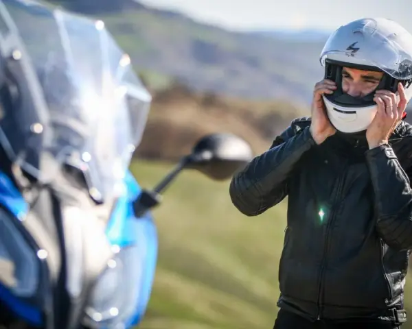 Uomo in giacca di pelle che indossa caschi per moto bianchi, pronto per un viaggio su una moto blu sfocata.