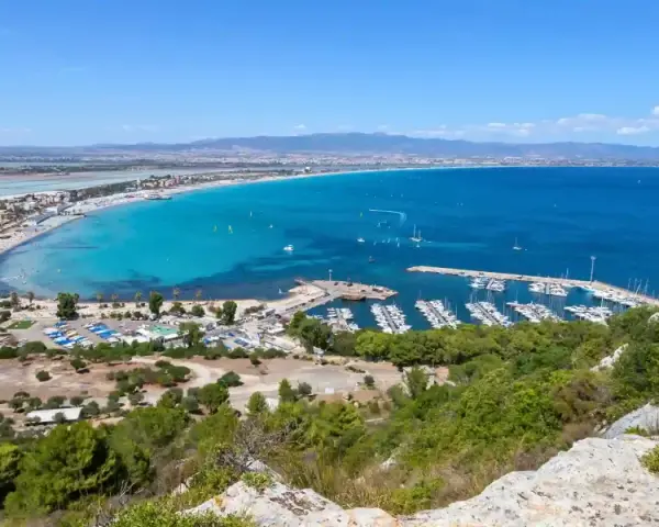 Vista panoramica della spiaggia del Poetto con mare turchese, porto turistico e saline rosa sullo sfondo.