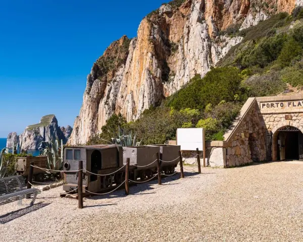 Antichi vagoni minerari e l'ingresso di Porto Flavia, testimoni della storica zona mineraria sarda, si stagliano contro un imponente promontorio roccioso.