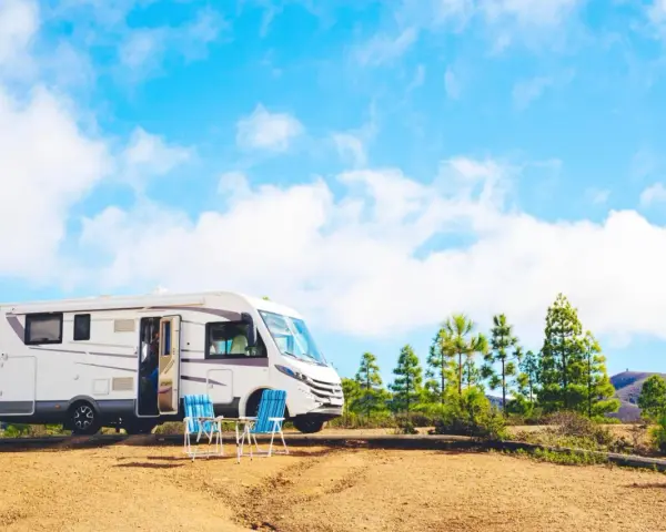 Un camper bianco è parcheggiato in un'area sterrata con due sedie blu e un tavolino. Sullo sfondo, pini e colline sotto un cielo azzurro con nuvole bianche. Un luogo ideale per un soggiorno in stile piscin'e janas.