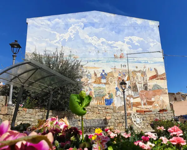 Un murale colorato a Loceri, Sardegna, raffigura scene di vita marina e rurale con persone, carri e animali sotto un cielo azzurro e nuvoloso.