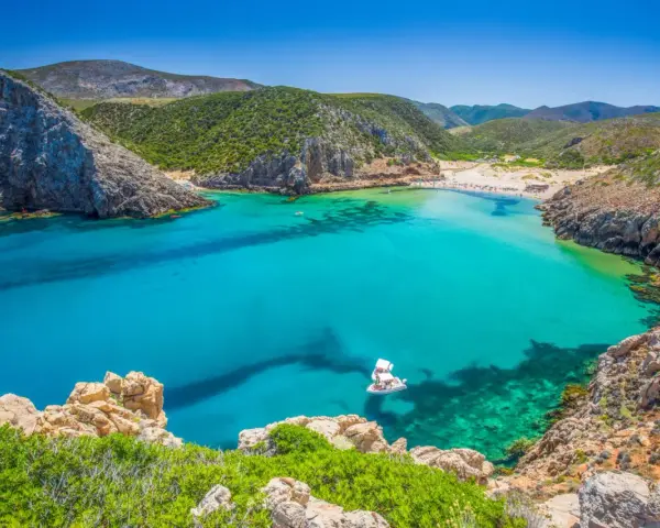 La splendida spiaggia di Buggerru, con acque cristalline e rocce imponenti, invita al relax. Una barca bianca solca il mare turchese.