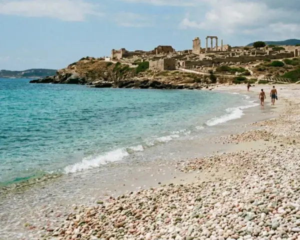Spiaggia di ciottoli con rovine antiche in collina e un faro in lontananza. Una coppia passeggia sulla riva, godendosi la brezza marina. La facella cabras illumina il paesaggio.