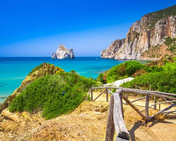 Sentiero con staccionata in legno conduce a una spiaggia con acqua cristallina e l'isolotto Pan di Zucchero in Sardegna.