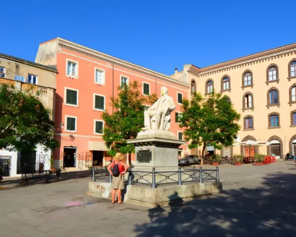 La piazza Tola a Sassari, con il monumento al centro, edifici colorati e alberi verdi sotto un cielo azzurro.