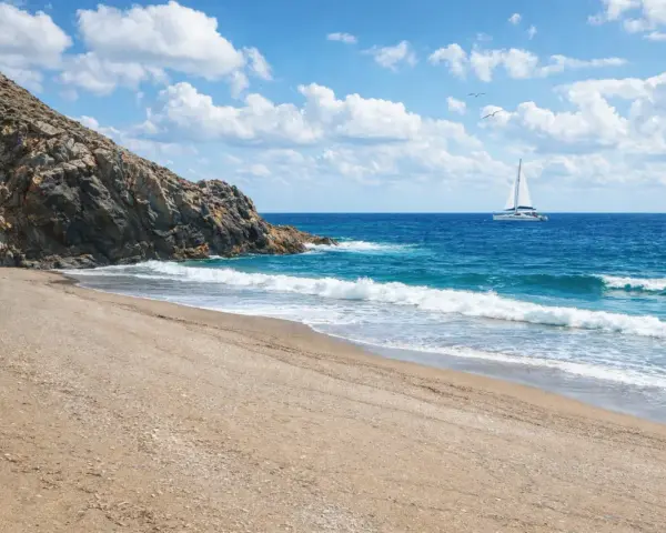 Spiaggia dorata e mare azzurro vicino a Sassari, con un catamarano bianco che naviga all'orizzonte sotto un cielo nuvoloso.
