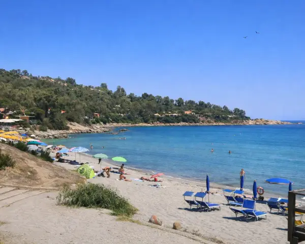 Spiaggia di Porto Frailis con ombrelloni colorati, lettini blu e gente che si gode il mare cristallino sotto il sole.
