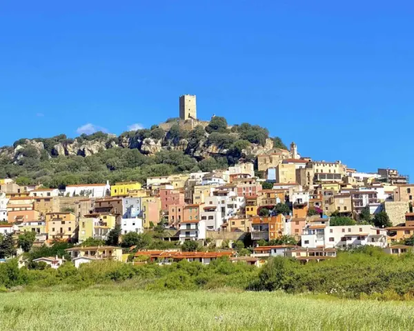 La **posada di sera** si staglia su una collina rocciosa, con case colorate e una torre medievale sotto un cielo azzurro.