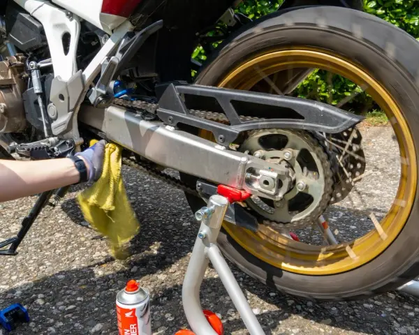 Mano con guanto pulisce catena moto con panno giallo. Spray pulitore catena e spazzola a terra.