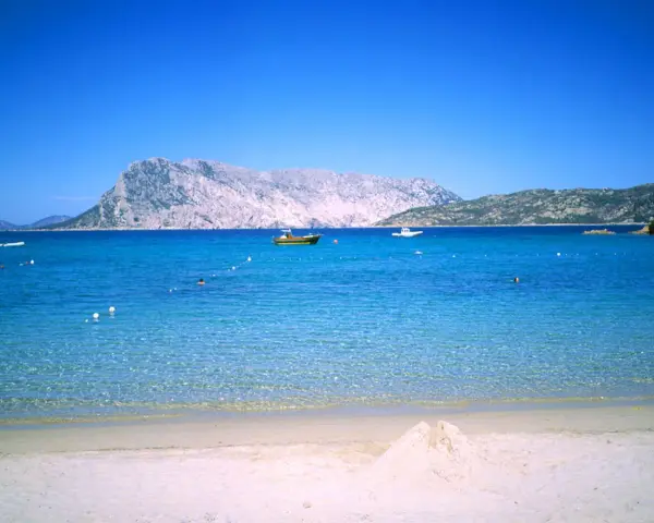 Spiaggia di Cala Suaraccia, mare cristallino, montagne sullo sfondo e barche in lontananza. Come arrivare: goditi il panorama!