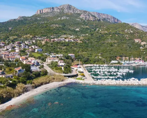 Vista aerea del centro storico di Santa Maria Navarrese, con case arroccate sulla collina, un porto turistico pieno di barche e il mare cristallino.