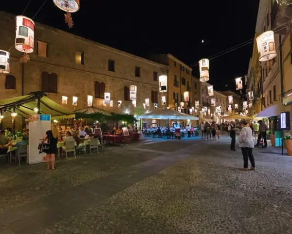 Serata ad Alghero: ristoranti all'aperto illuminati da lanterne, gente che passeggia e cena.