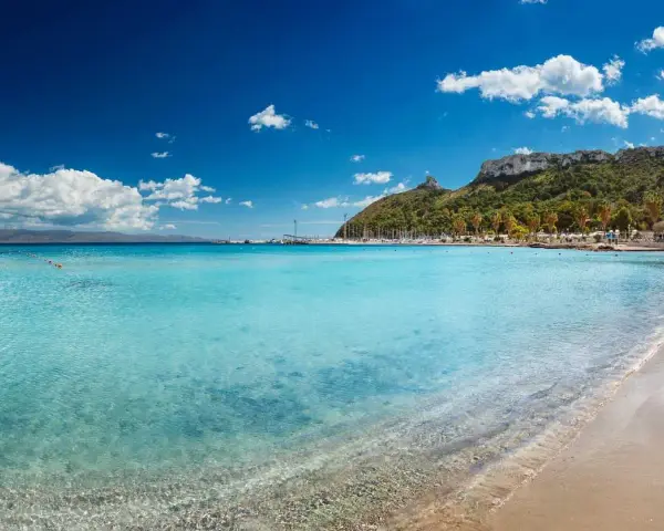Acque cristalline e sabbia dorata sulle spiagge di Cagliari, con il promontorio verdeggiante e il porto turistico sullo sfondo.