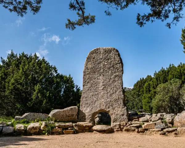 La tomba dei giganti in Sardegna, un imponente monolito di granito con un arco, circondato da pietre antiche e vegetazione rigogliosa sotto un cielo azzurro.