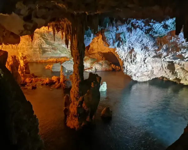 Interno di una grotta con stalattiti e stalagmiti illuminate da luci arancioni e blu. L'acqua scura riflette le formazioni rocciose in questa grotta verde.