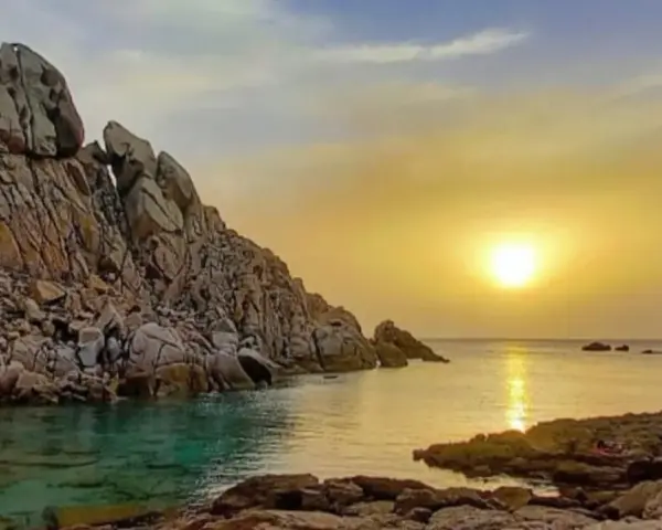 Scogliere rocciose bagnate da acque cristalline, con il sole che tramonta sulla valle della luna, creando riflessi dorati sul mare.