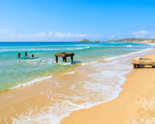 Spiaggia di Chia, Cagliari. Resti di antiche strutture in mare e sulla sabbia dorata, con acque turchesi e cielo azzurro.