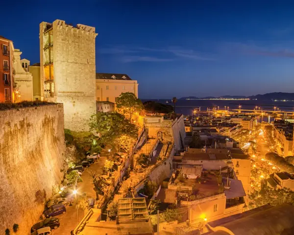 Torre medievale e palazzi illuminati nella piazza di Cagliari, con il porto sullo sfondo al crepuscolo.