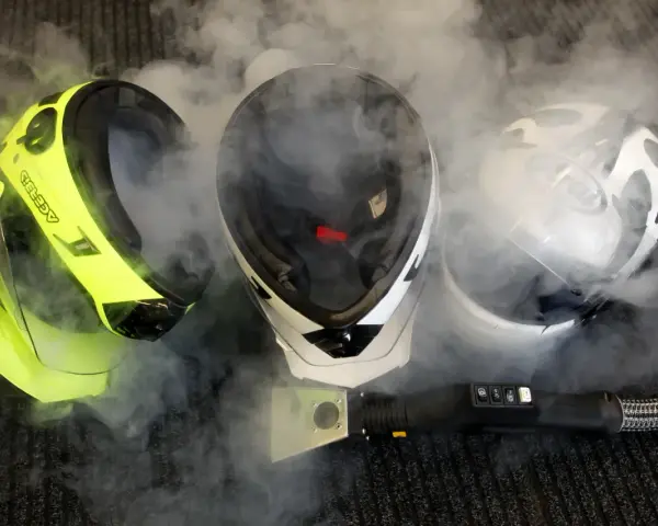 Tre caschi, uno giallo fluo, uno bianco e nero, e uno bianco, sono avvolti dal vapore. Un accessorio per la pulizia a vapore è posizionato davanti a loro, suggerendo che il casco non deve essere pulito con solventi.