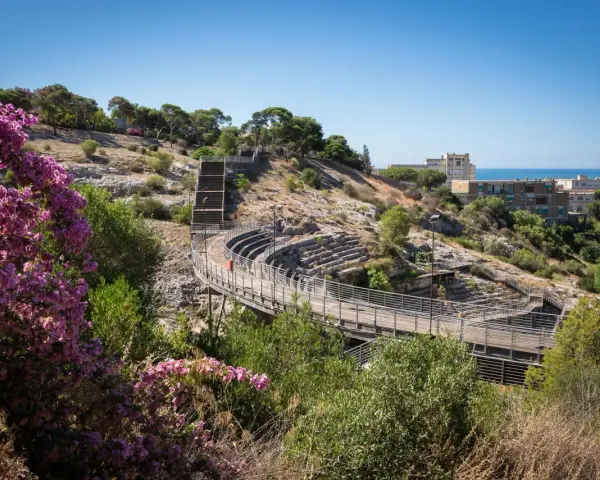 Un suggestivo anfiteatro a Cagliari, con gradinate in pietra e passerelle in legno, immerso nel verde e con fiori viola in primo piano.