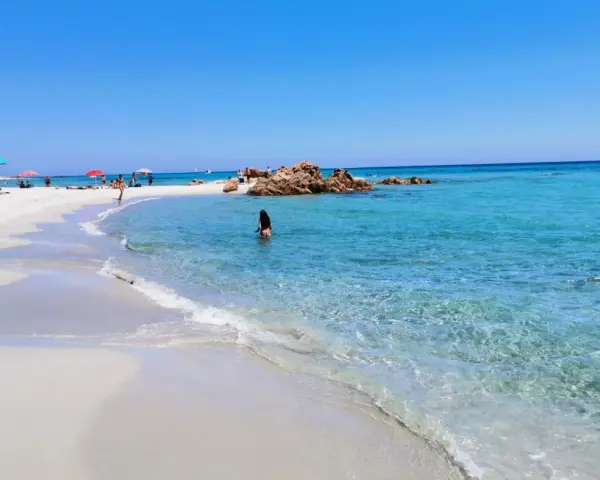 Spiaggia di Berchida: sabbia bianca, acque cristalline e rocce rosse sotto un cielo azzurro. Una donna si gode il mare calmo.