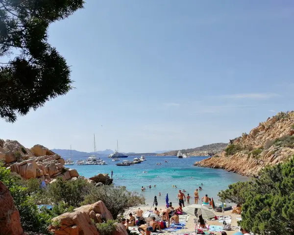 Spiaggia affollata a Cala Coticcio, con gente che prende il sole e nuota nelle acque cristalline. Diverse barche e yacht ancorati nella baia.