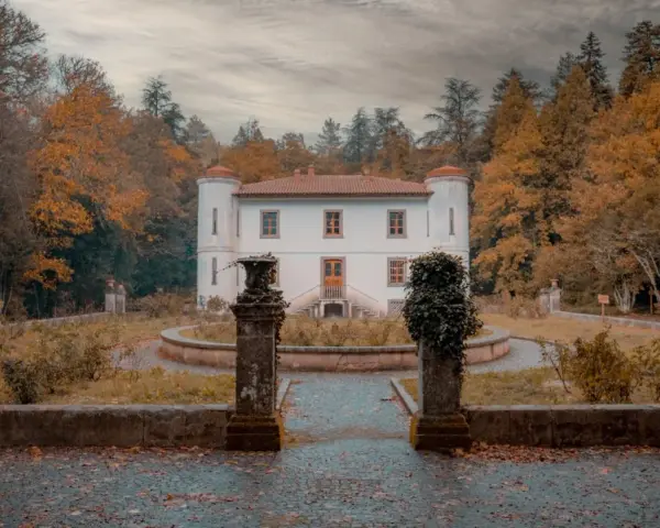 Un'antica villa bianca con torrette, circondata da alberi autunnali dai colori caldi. Un sentiero lastricato conduce all'ingresso, tra statue e vegetazione. Le **badde salighes** sussurrano storie di tempi passati.