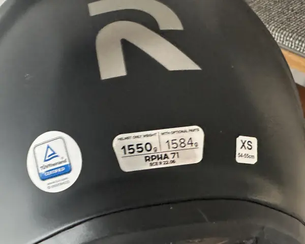 Casco nero con logo R argentato. Etichette indicano peso, modello RPHA 71, certificazione e taglia XS. Sul casco possono essere applicati adesivi.