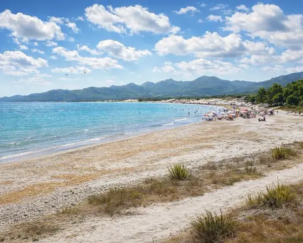 Spiaggia affollata con ombrelloni e gente che si gode il sole. Il nuoro mare azzurro lambisce la costa, con montagne verdi sullo sfondo.