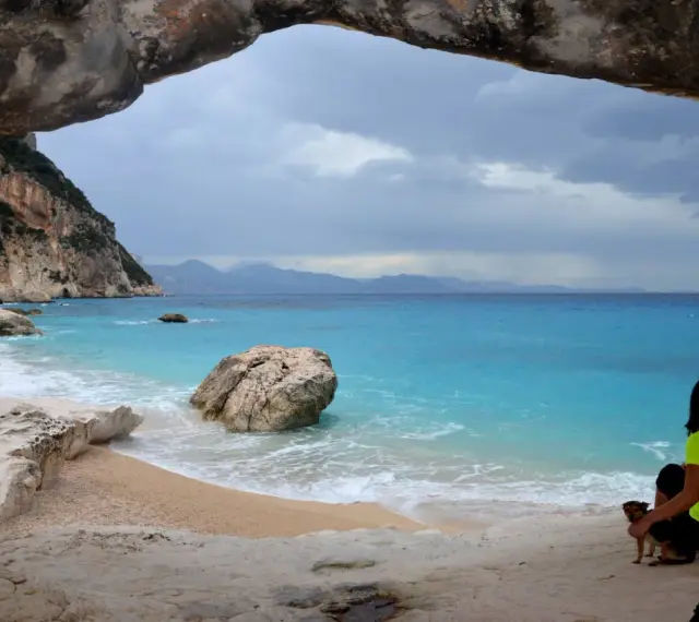 Vista da una grotta su Cala Goloritzé, con mare turchese, scogliere e una donna con un cane.