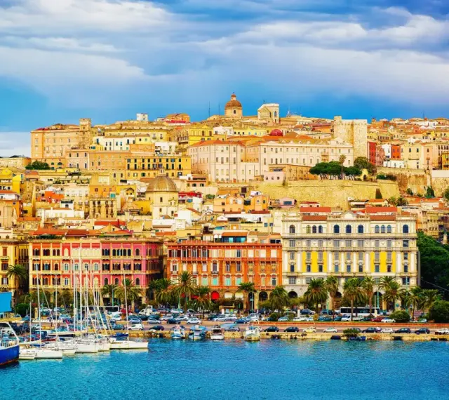 Panoramica colorata di **Mirrionis**, con edifici storici, un porto con barche a vela e un cielo azzurro con nuvole.