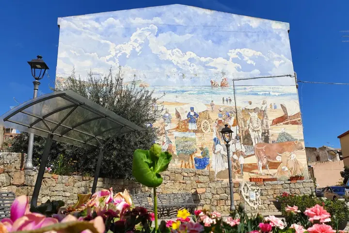 Un murale colorato a Loceri, Sardegna, raffigura scene di vita marina e rurale con persone, carri e animali sotto un cielo azzurro e nuvoloso.