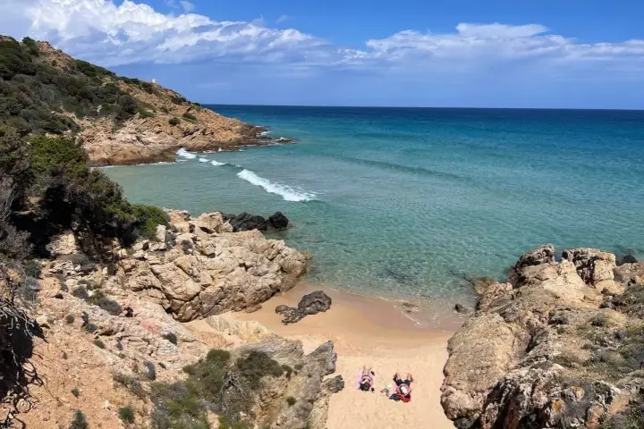 Spiaggia selvaggia vicino al centro storico di Chia, con rocce dorate, mare cristallino e due persone che prendono il sole.