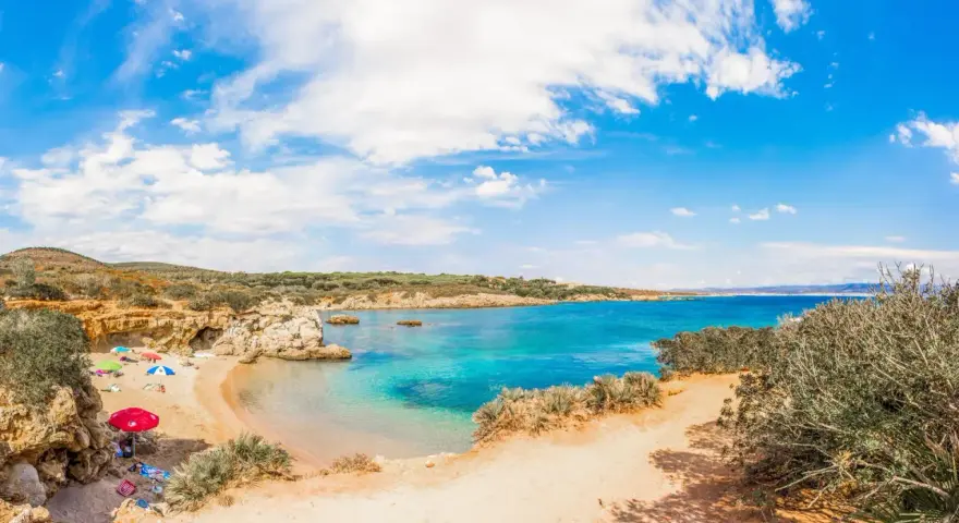 Spiaggia Lazzaretto ad Alghero: sabbia dorata, acque cristalline e macchia mediterranea sotto un cielo azzurro con nuvole bianche.