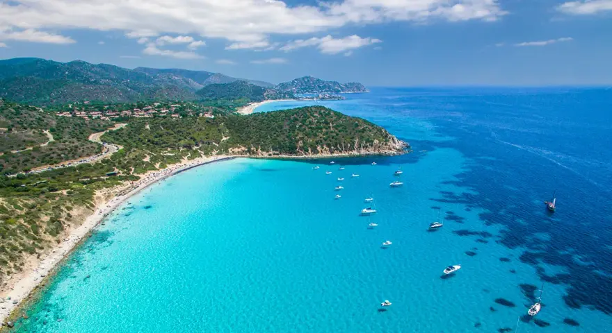 Spiaggia di Chia, Sardegna: acque cristalline che sembrano mari pintau, con barche a vela e un litorale affollato.