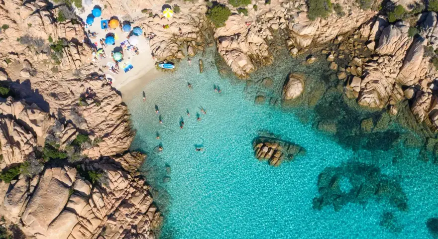 Vista aerea della spiaggia di Cala Coticcio, con bagnanti che si godono le acque cristalline e ombrelloni colorati sulla sabbia dorata.