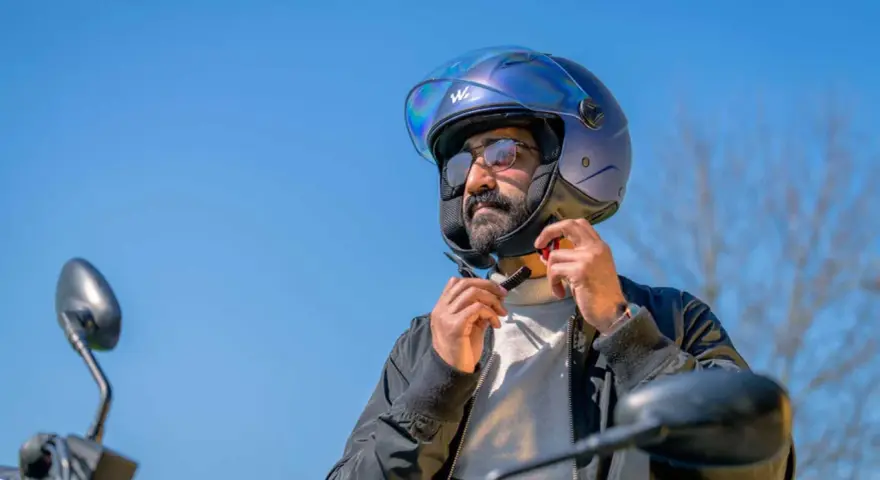 Uomo con barba e occhiali da sole che regola il cinturino di un casco da moto blu iridescente. La corretta misura casco moto è fondamentale per la sicurezza.