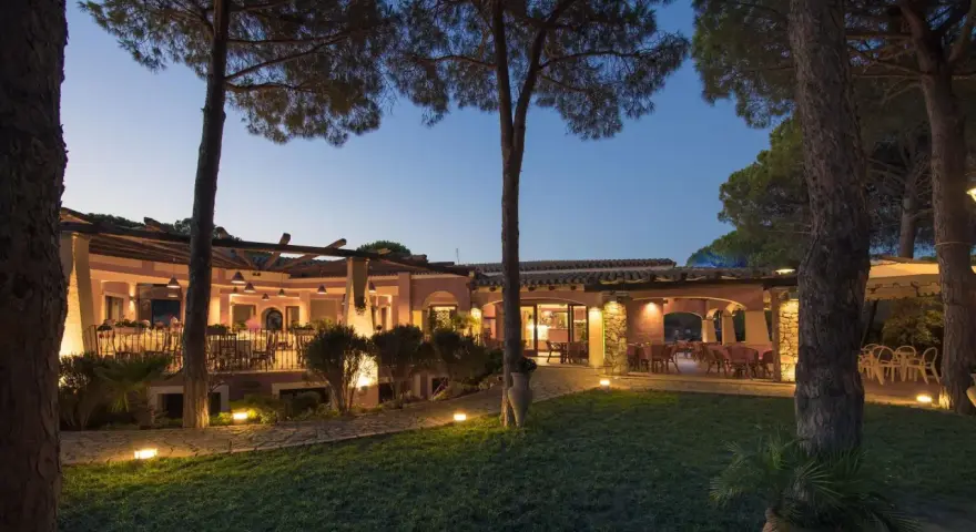 La sorgente Villasimius: un ristorante con tavoli all'aperto illuminati da luci soffuse, circondato da pini e un prato curato.