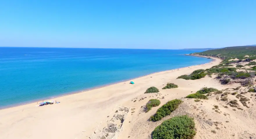 Vasta spiaggia di sabbia dorata, mare azzurro e vegetazione mediterranea sulla spiaggia di Piscinas. Rovine antiche emergono dalla sabbia.