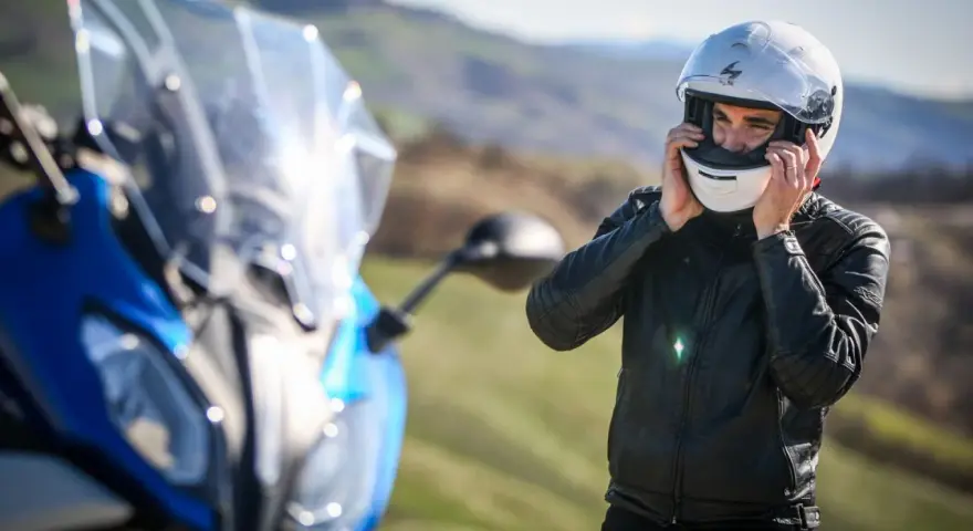 Uomo in giacca di pelle che indossa caschi per moto bianchi, pronto per un viaggio su una moto blu sfocata.
