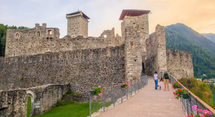 Un sentiero fiorito conduce al maestoso Castello San Michele, con le sue torri imponenti e mura antiche che si stagliano contro il cielo.
