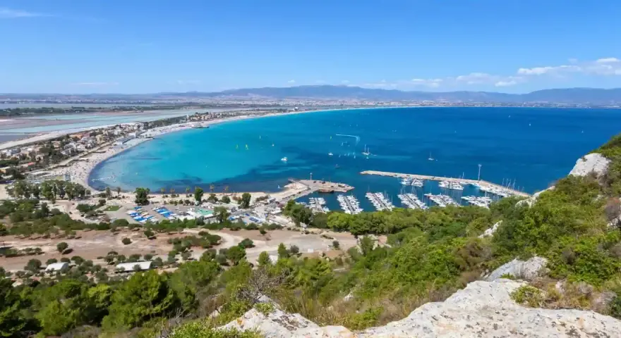 Vista panoramica della spiaggia del Poetto con mare turchese, porto turistico e saline rosa sullo sfondo.