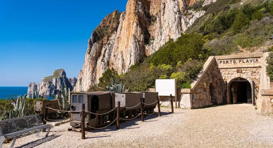 Antichi vagoni minerari e l'ingresso di Porto Flavia, testimoni della storica zona mineraria sarda, si stagliano contro un imponente promontorio roccioso.
