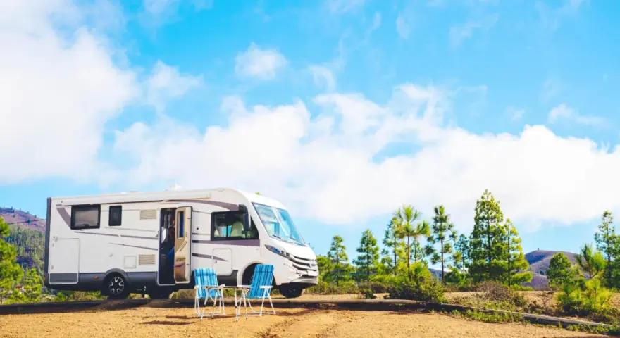 Un camper bianco è parcheggiato in un'area sterrata con due sedie blu e un tavolino. Sullo sfondo, pini e colline sotto un cielo azzurro con nuvole bianche. Un luogo ideale per un soggiorno in stile piscin'e janas.