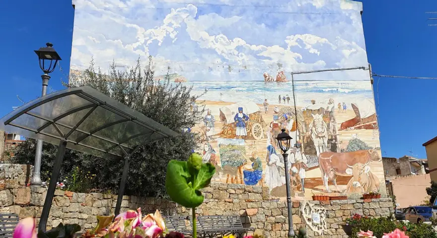 Un murale colorato a Loceri, Sardegna, raffigura scene di vita marina e rurale con persone, carri e animali sotto un cielo azzurro e nuvoloso.