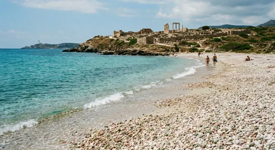 Spiaggia di ciottoli con rovine antiche in collina e un faro in lontananza. Una coppia passeggia sulla riva, godendosi la brezza marina. La facella cabras illumina il paesaggio.