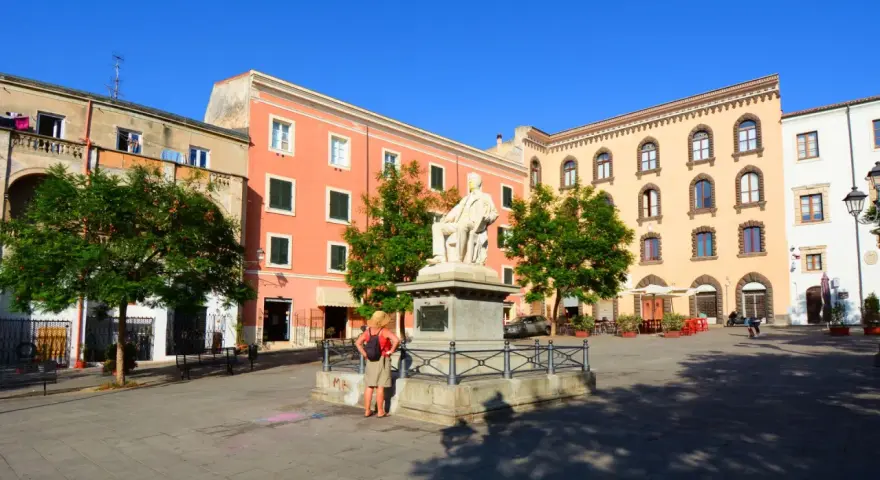 La piazza Tola a Sassari, con il monumento al centro, edifici colorati e alberi verdi sotto un cielo azzurro.